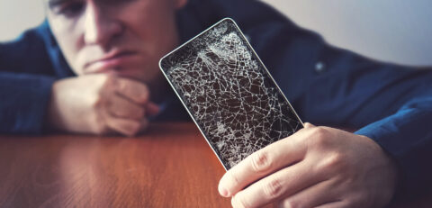 Hands holding a mobile phone with a broken screen over the wooden surface. A man looks at his broken phone lying on a wooden table with a sad look. guy was upset about breaking his phone.